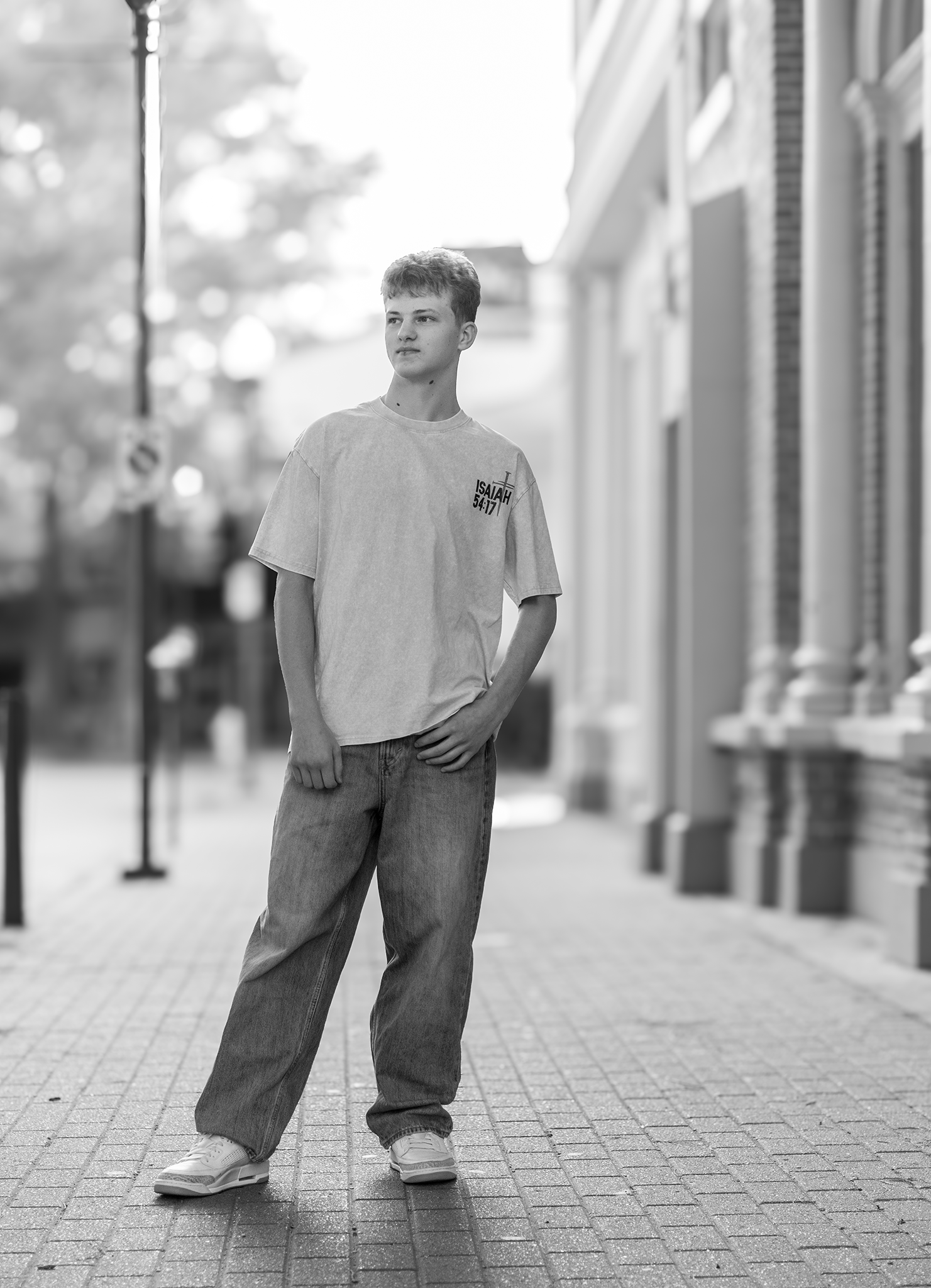 Black and white photo of teenage boy in oversized tee and jeans on a city sidewalk.