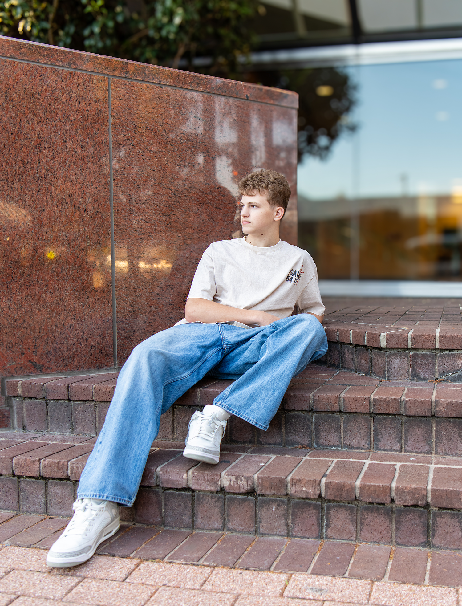 Teen boy in white tee and jeans sitting on brick steps outside a modern building.
