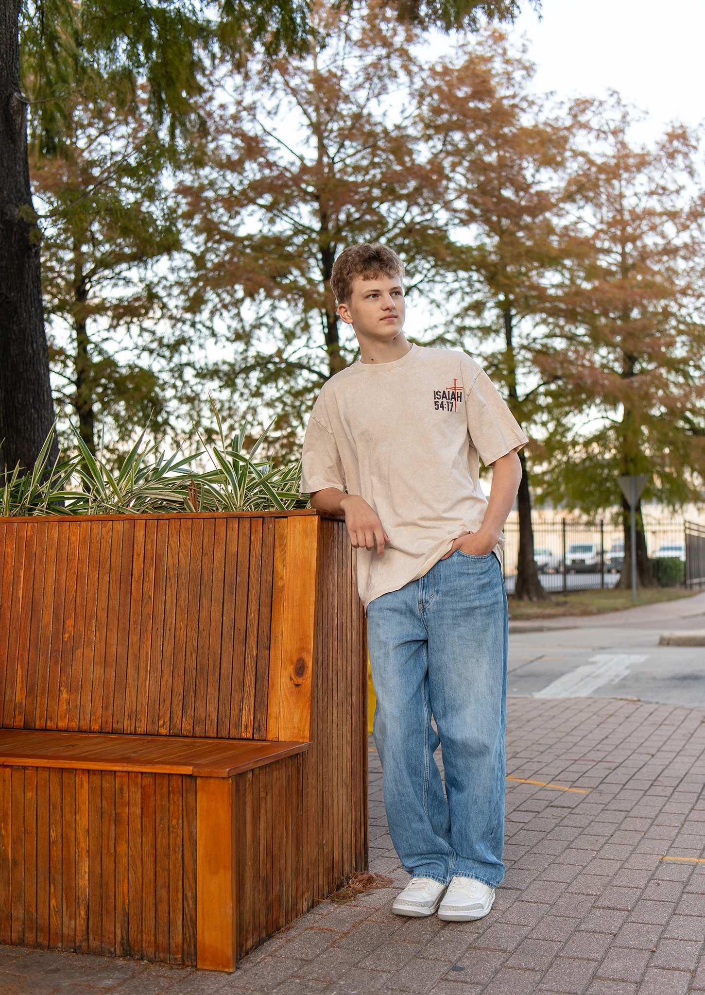 Young man in beige Isaiah 54:17 tee and wide-leg jeans leaning on wooden planter box outdoors.