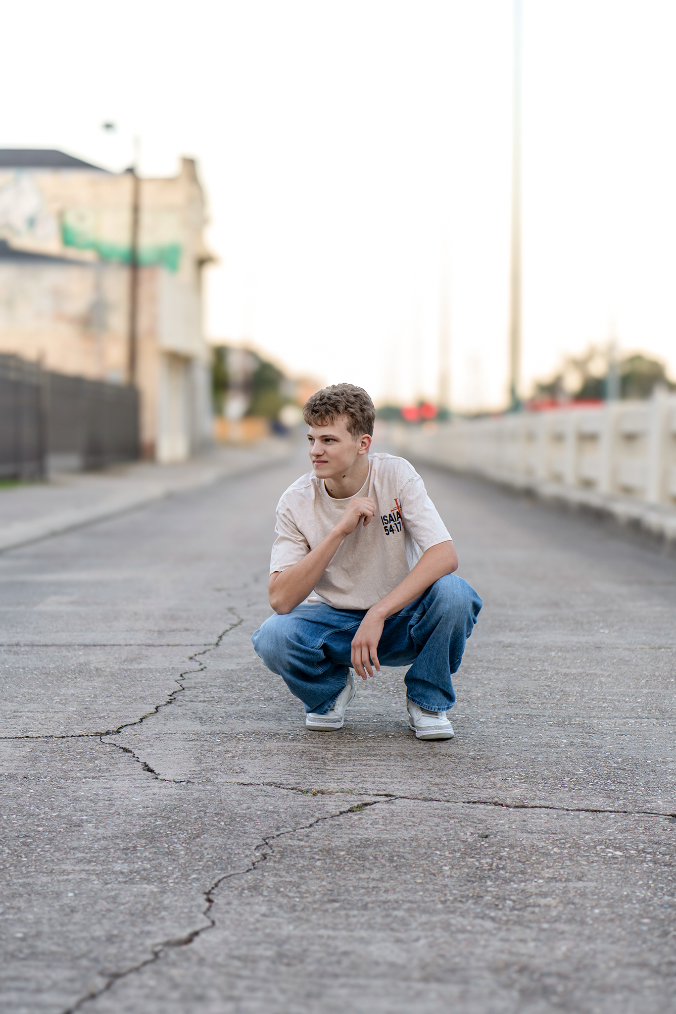 Young man in white tee and baggy jeans crouching on a cracked urban road during golden hour.