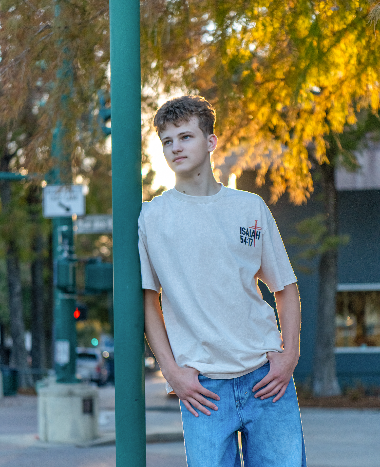 Teen boy in beige Isaiah 54:17 graphic tee and jeans leaning on green pole on city street.