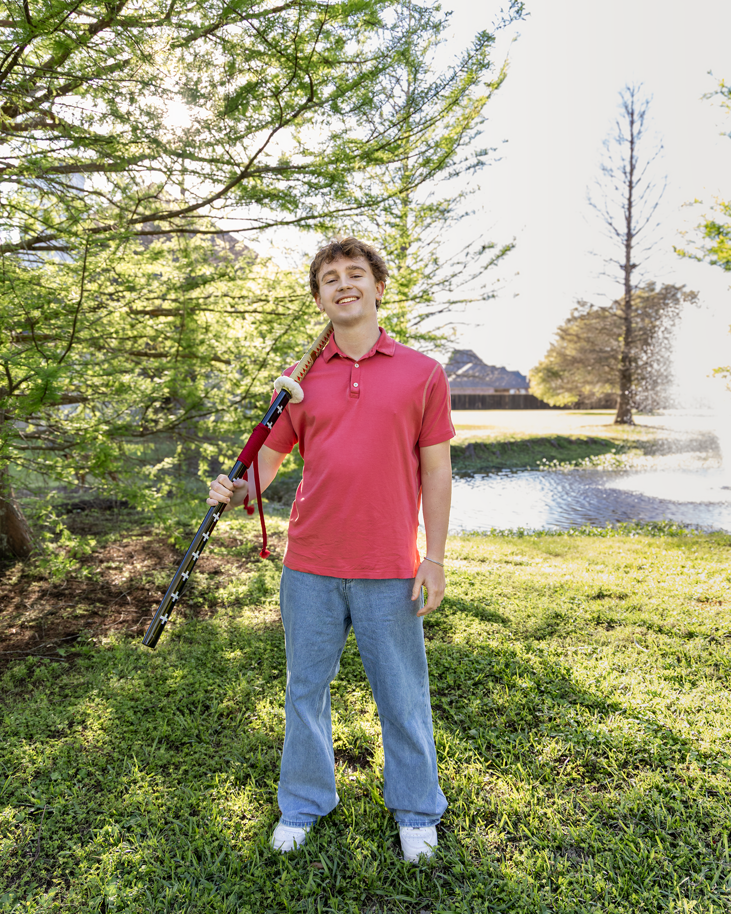 Smiling teen in red polo holds clarinet while standing on sunny green lawn.