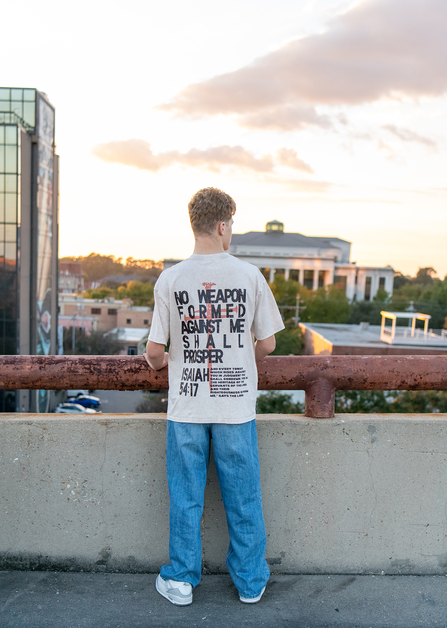 Back view of man wearing Isaiah 54:17 graphic tee on rooftop with city skyline at sunset.