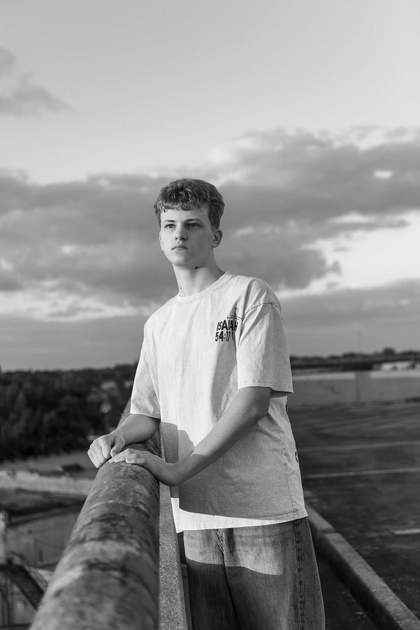 Black and white portrait of teen in graphic tee leaning on railing with cloudy sky backdrop.