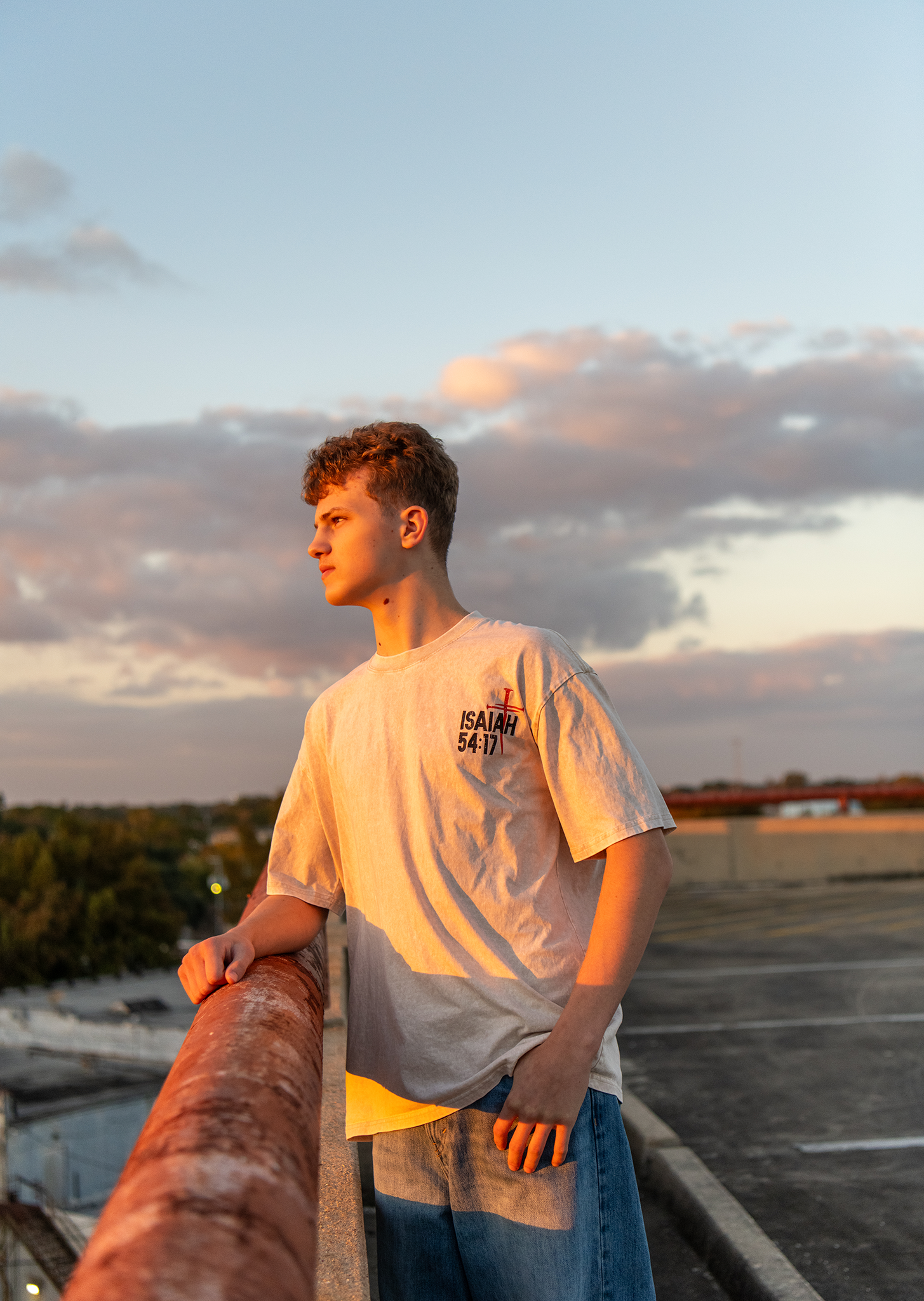 Young man in Isaiah 54:17 graphic tee leaning on rusty railing at golden hour on rooftop.
