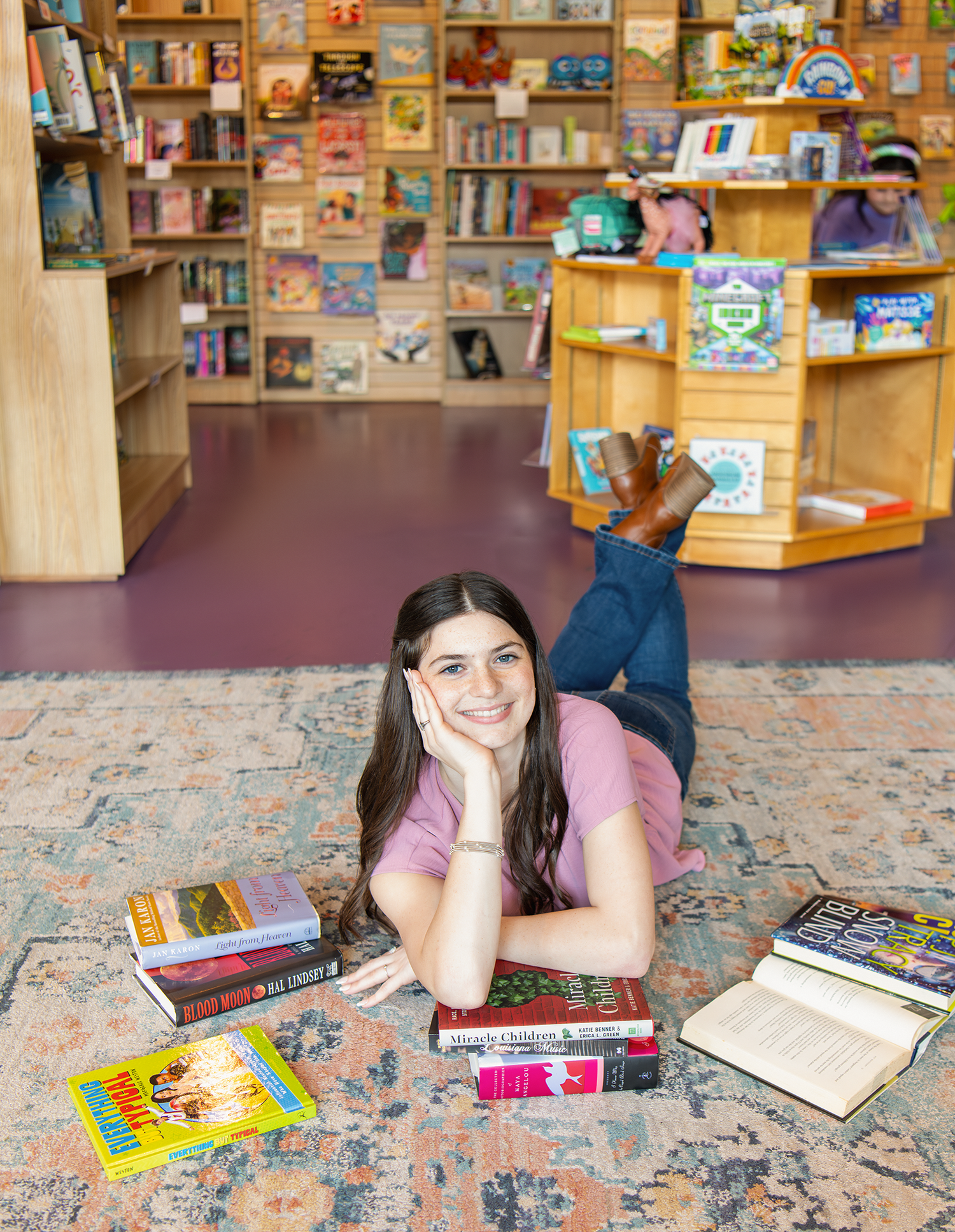 Smiling teen girl lying on bookstore floor surrounded by books in a pink top and jeans.