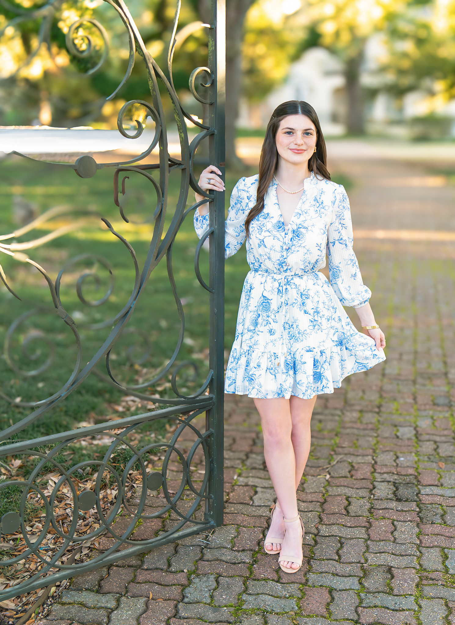 Teen girl in blue floral dress posing by ornate iron gate on brick path in sunny park.