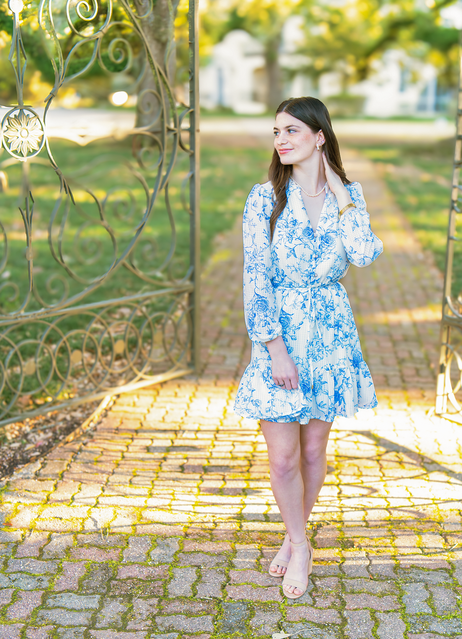 Teen girl in blue floral dress walking through iron gate on golden brick path in park.