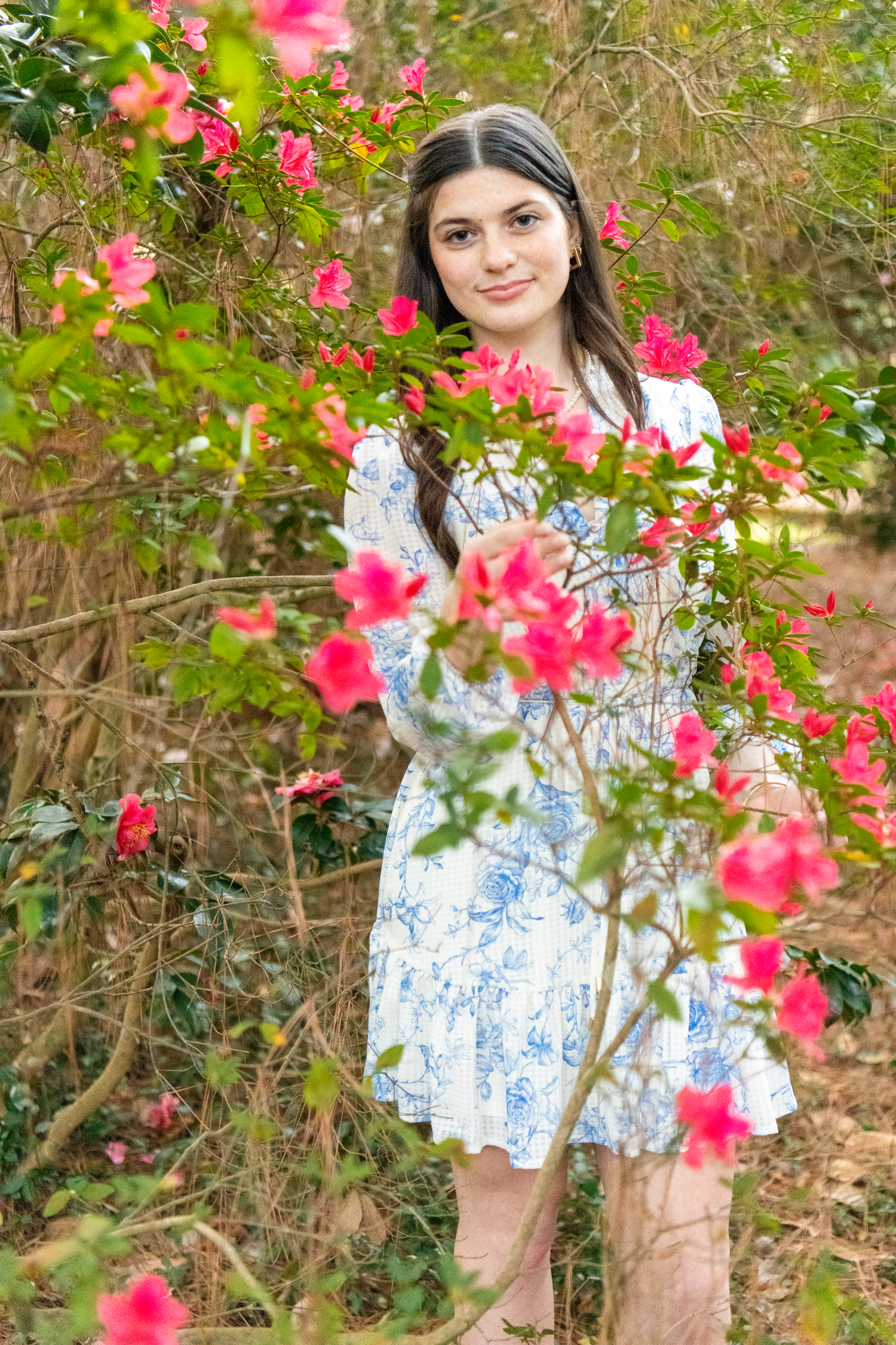 Teen girl in blue floral dress smiling among vibrant pink azalea blooms in a garden.