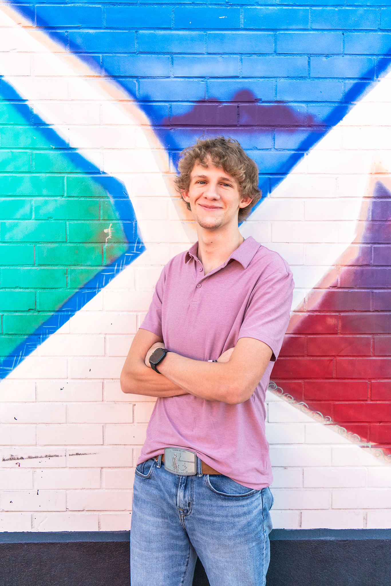 Smiling teen boy in pink polo with arms crossed in front of colorful mural wall.