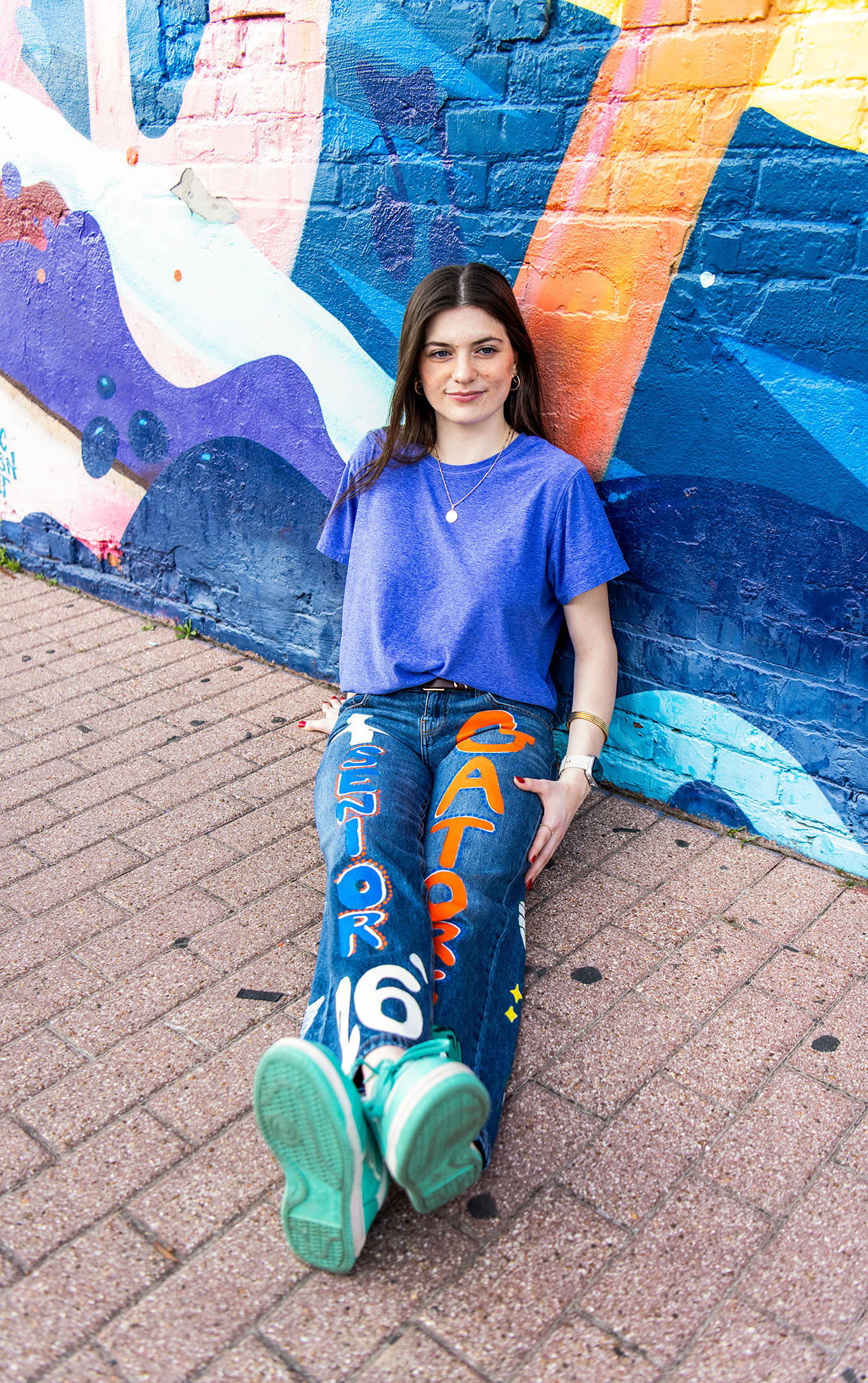Young woman in blue tee sitting against vibrant mural wearing painted Gators jeans.