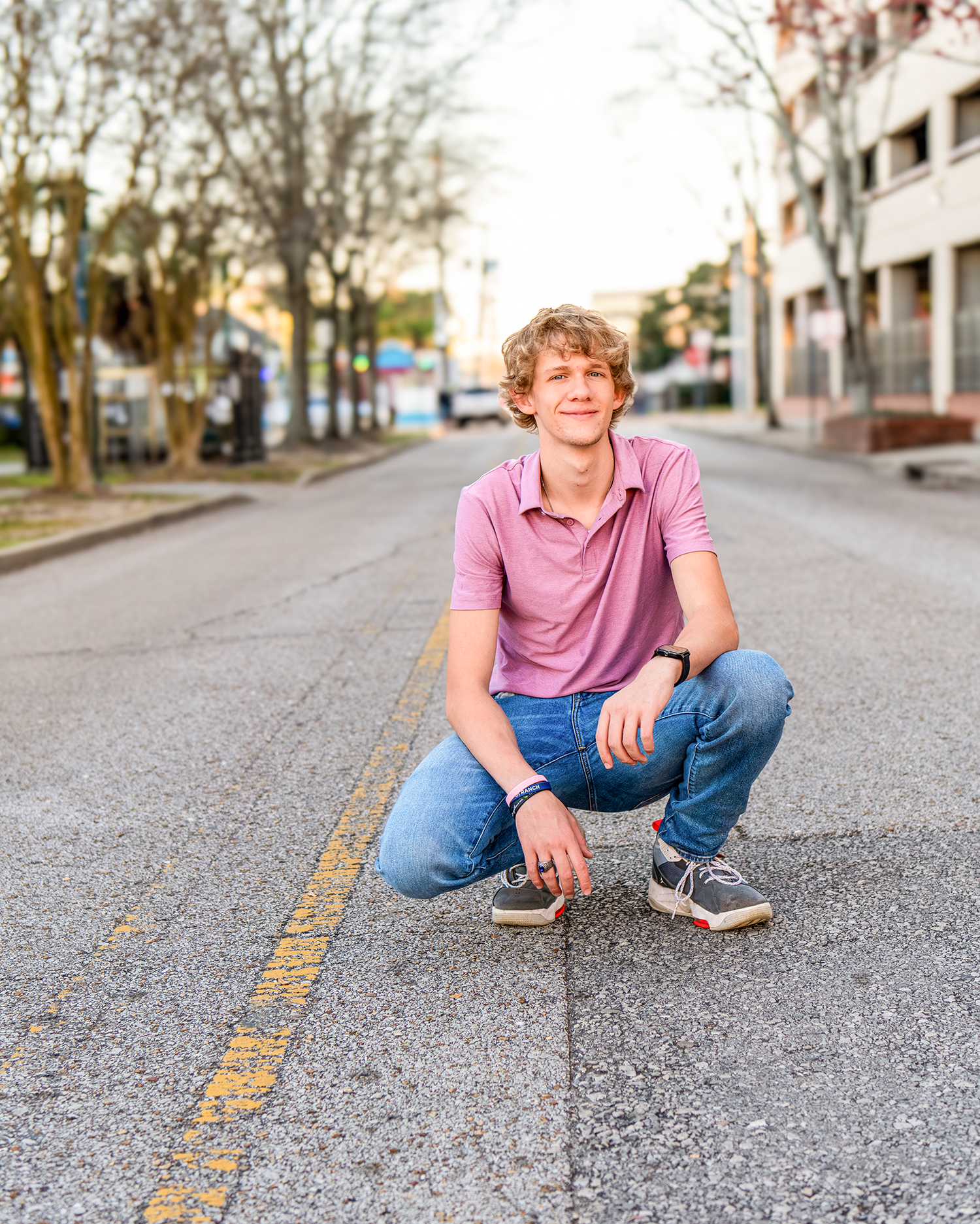 Young man in pink polo and jeans crouching on a urban street for a senior portrait photo.