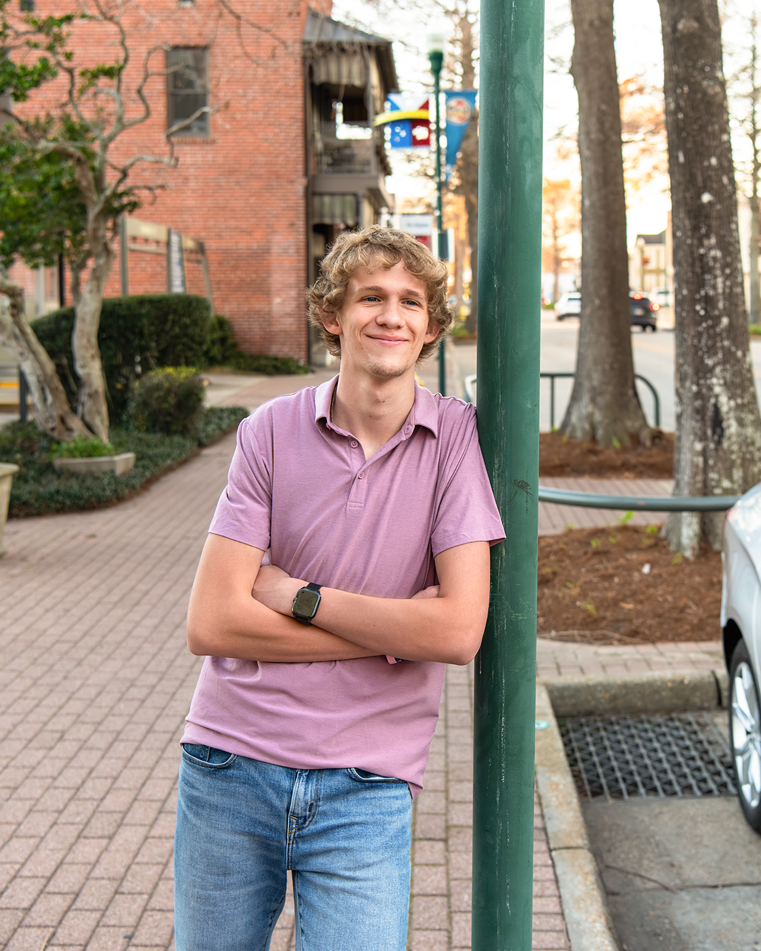 Smiling teen boy in pink polo leaning on green pole along a tree-lined sidewalk.