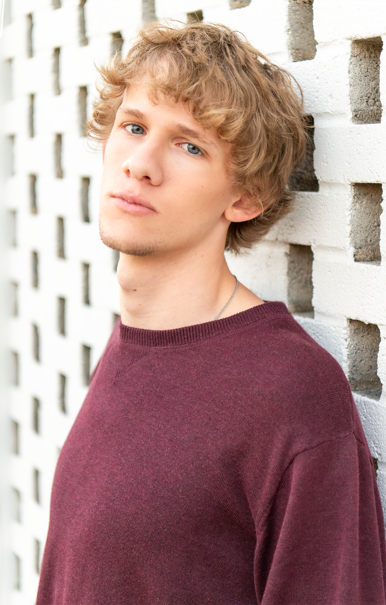 Close-up portrait of teen boy in maroon sweater leaning against white lattice brick wall.