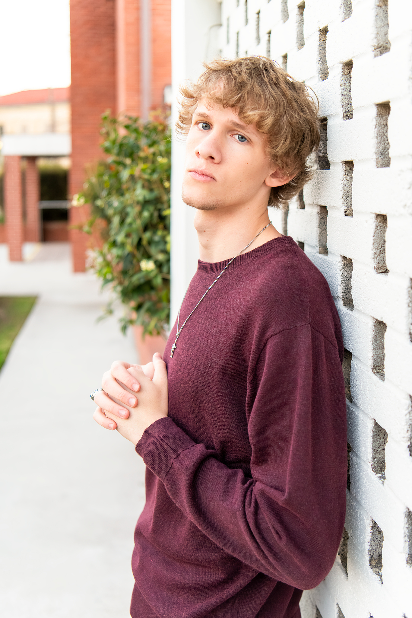 Teen boy in maroon sweater leaning against white brick wall for a senior portrait photo.