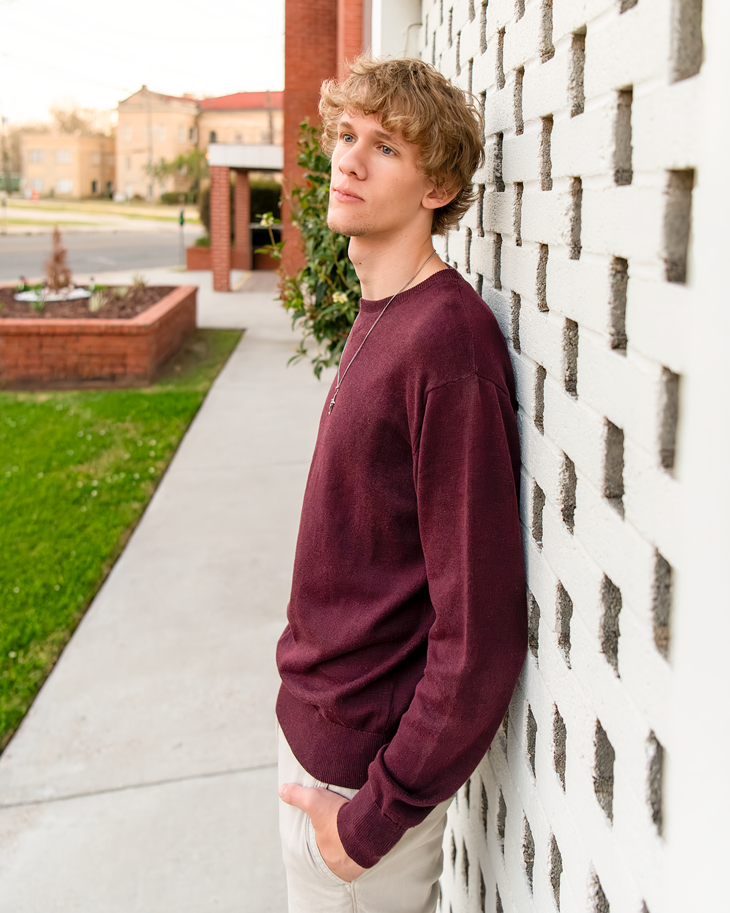 Teen boy in burgundy sweater leaning against white brick wall for an outdoor senior portrait.