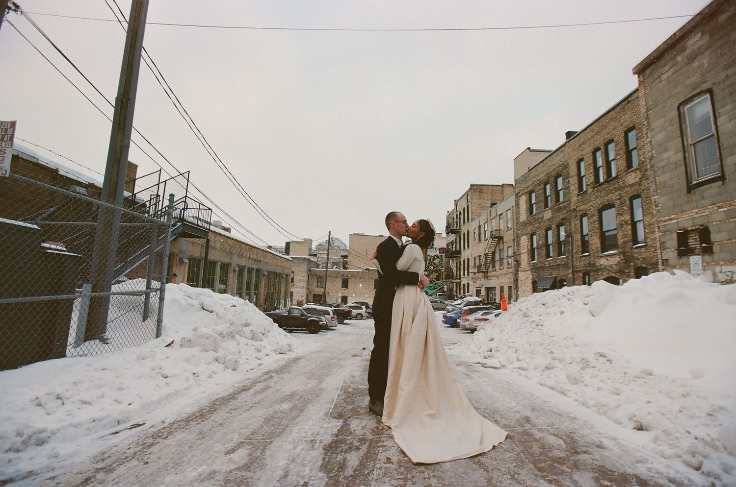 A winter wedding photoshoot on a snow-covered city street with brick buildings.