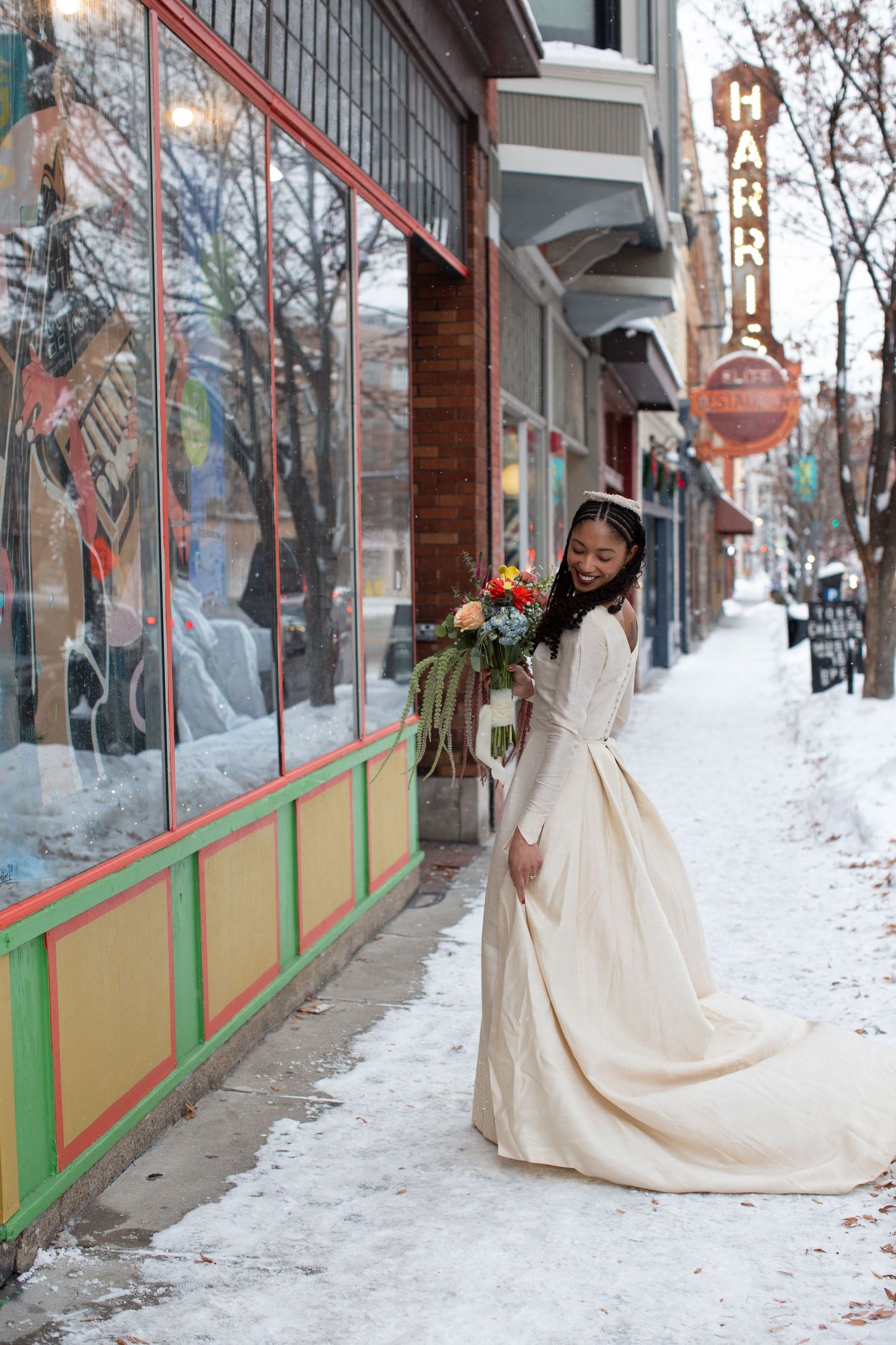 A bride in a flowing white dress twirls playfully in the snow outside storefronts.