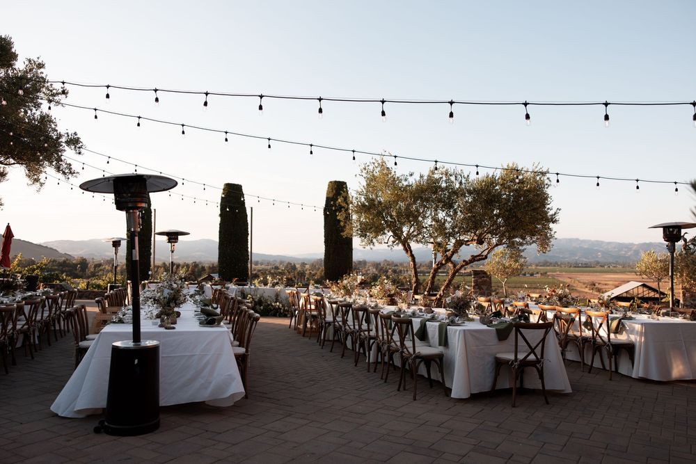 Outdoor wedding reception setup with string lights and heaters overlooking scenic mountain views at sunset.