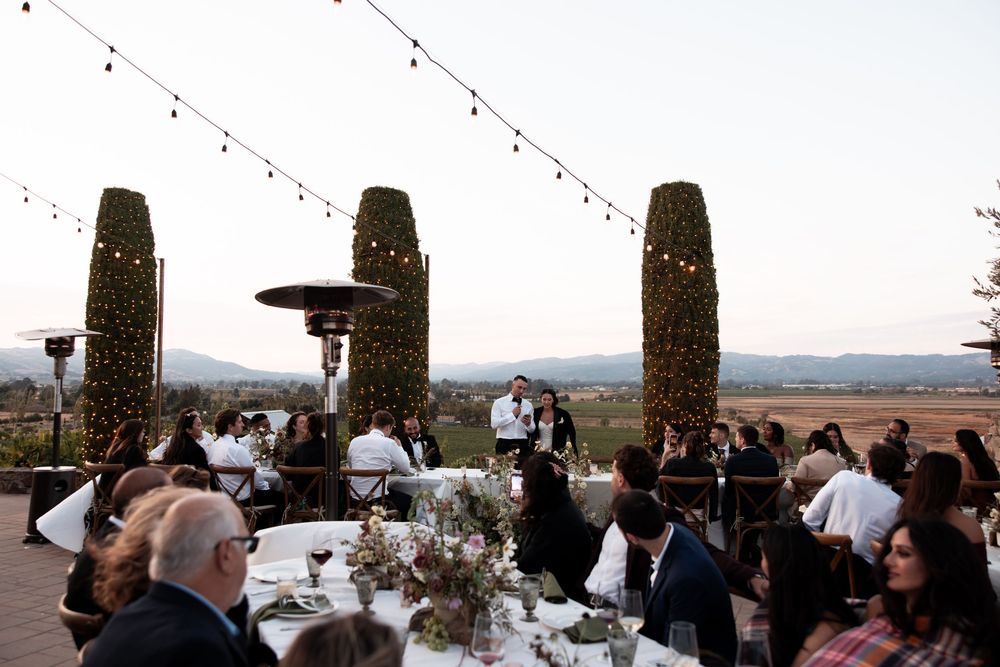 Outdoor wedding reception at sunset with string lights and tall stone pillars overlooking a mountain landscape.