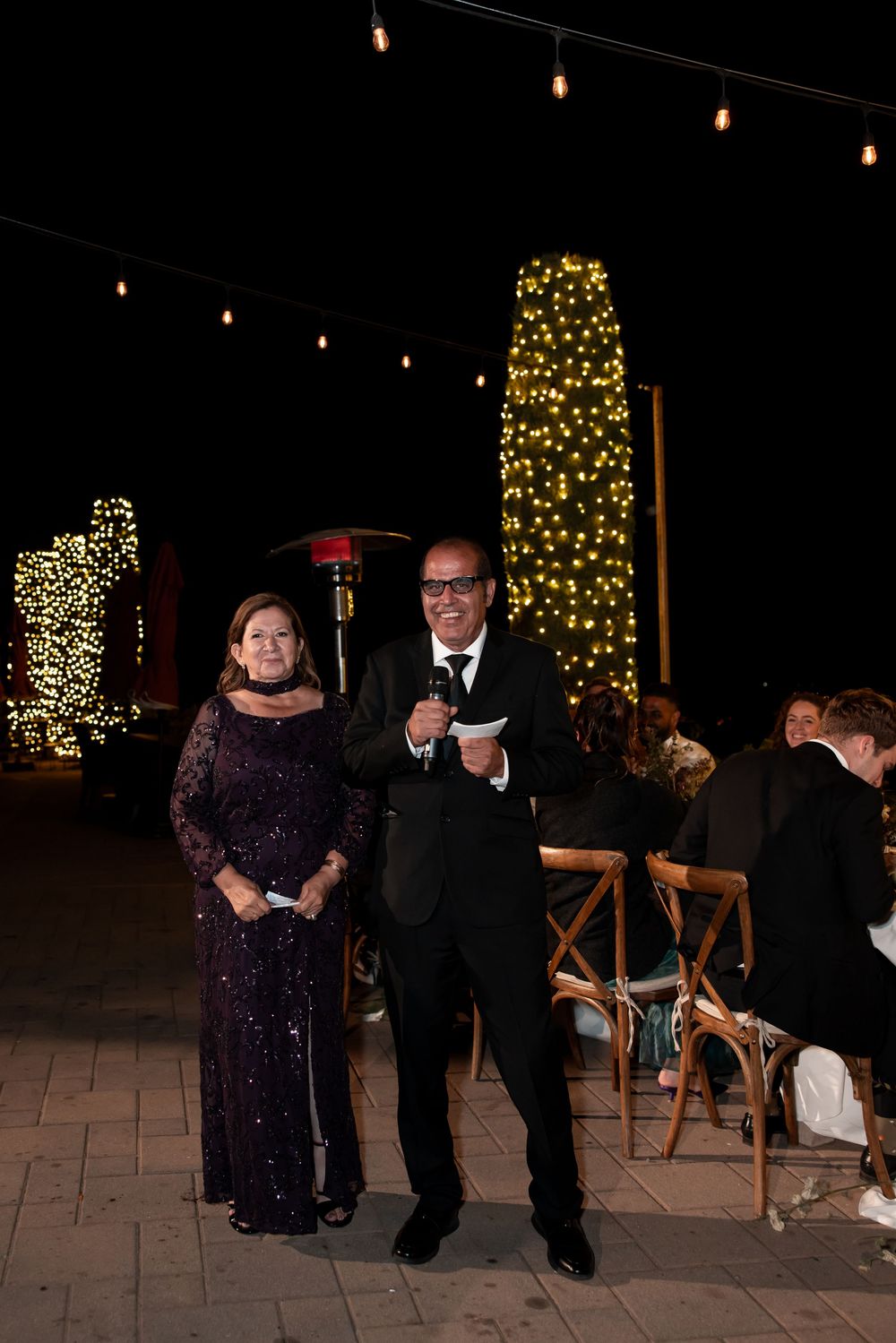 Couple dancing at an evening celebration with illuminated Christmas trees and string lights in the background.