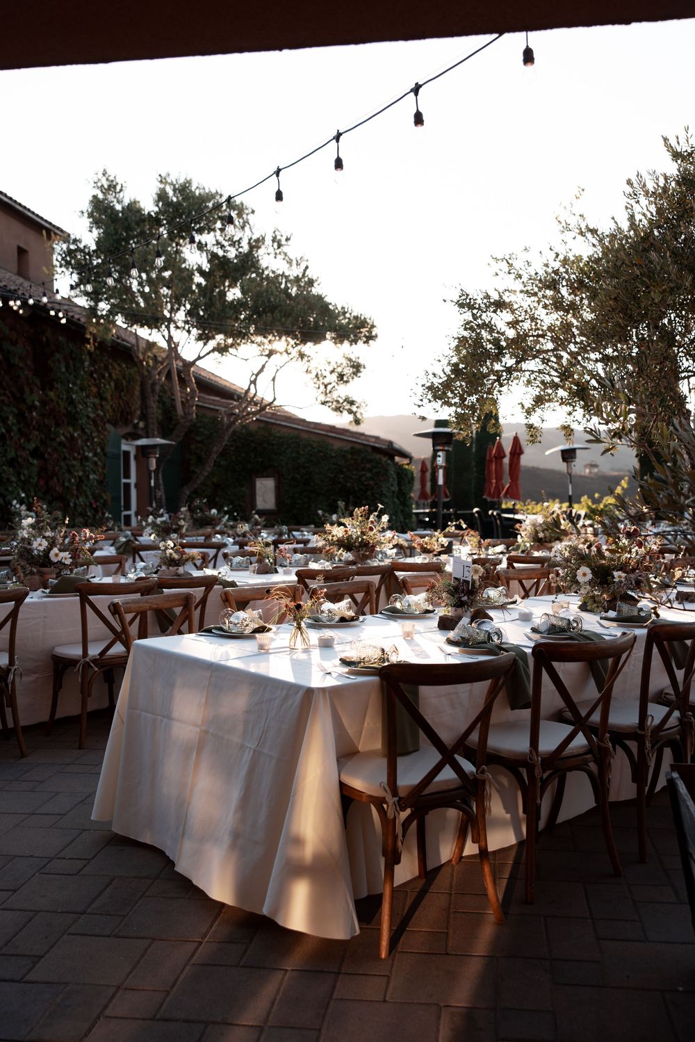 Elegant outdoor wedding reception tables with white linens and string lights at dusk create a romantic ambiance.