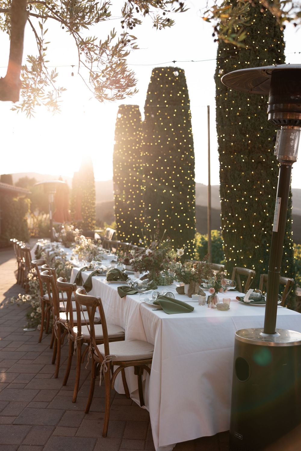 Elegant outdoor dining setup with white tablecloths and cypress trees at sunset, illuminated by string lights above.