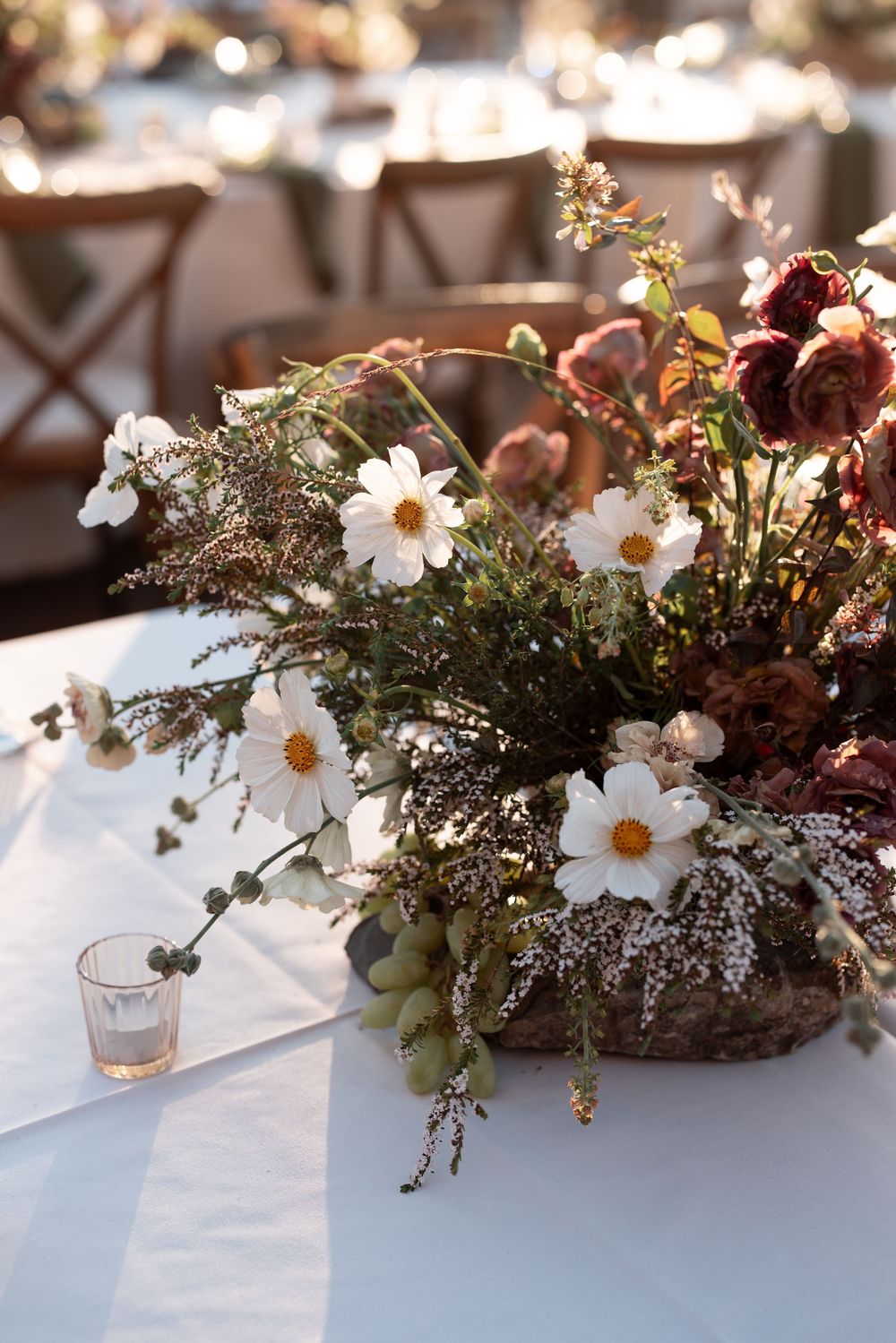 Rustic floral centerpiece with white daisies and greenery on a white tablecloth in warm sunlight.
