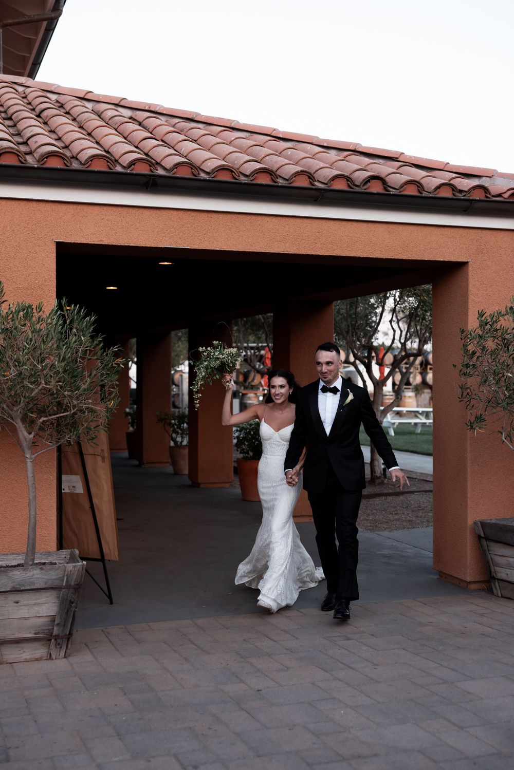A wedding couple walks under a covered terrace with Spanish-style terracotta roof tiles and coral-colored walls.