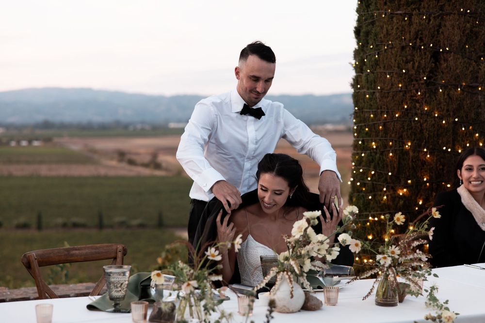A romantic outdoor wedding reception table setting at dusk with string lights and rustic decor in a vineyard.