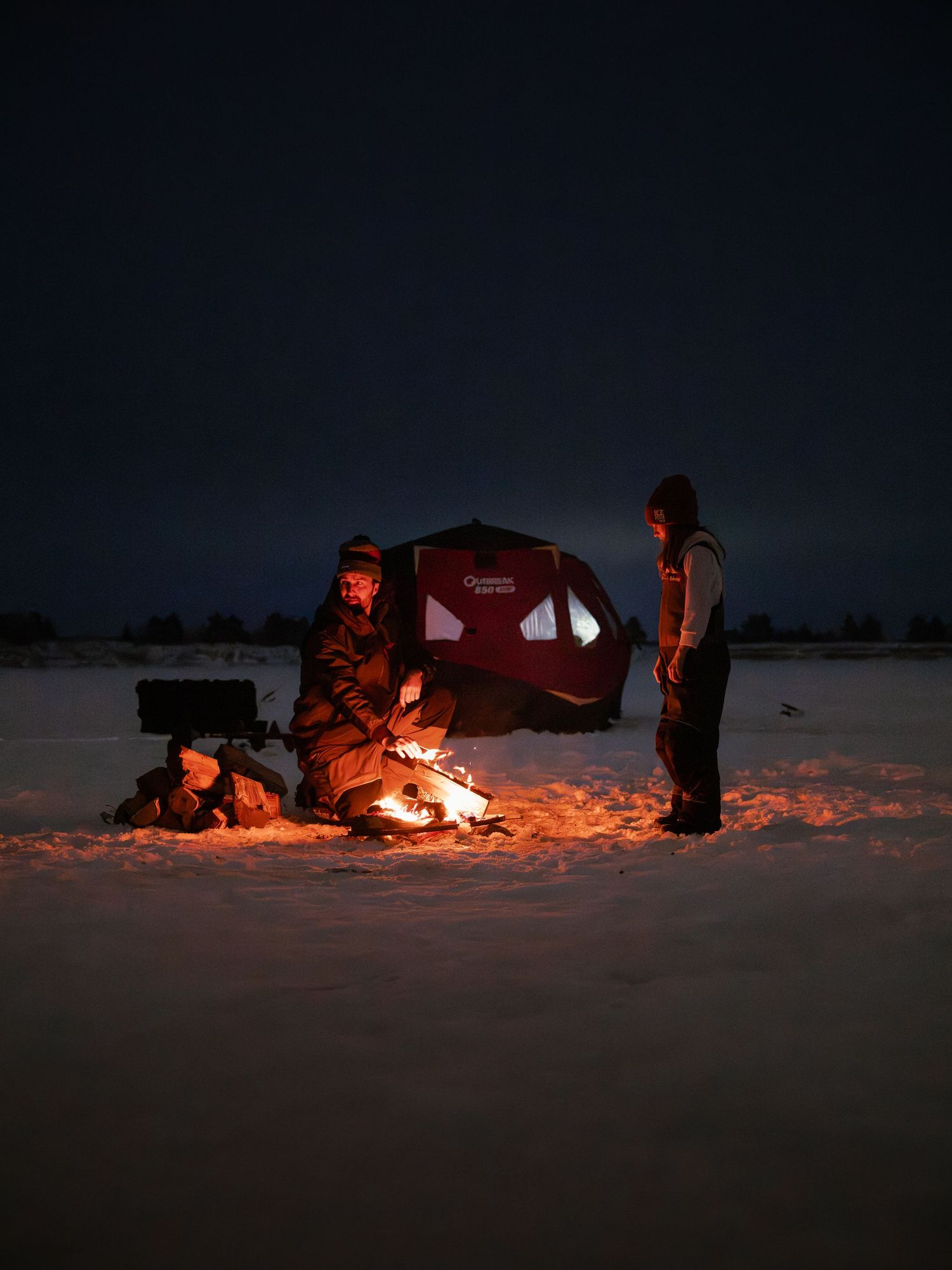 Ice fishing at night with red tent and campfire glowing on snowy lake.