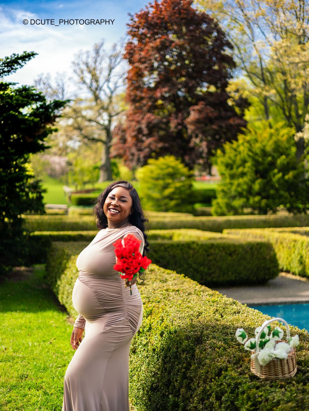 Smiling pregnant woman in beige gown holding red flowers in a lush garden park during a maternity photo shoot.