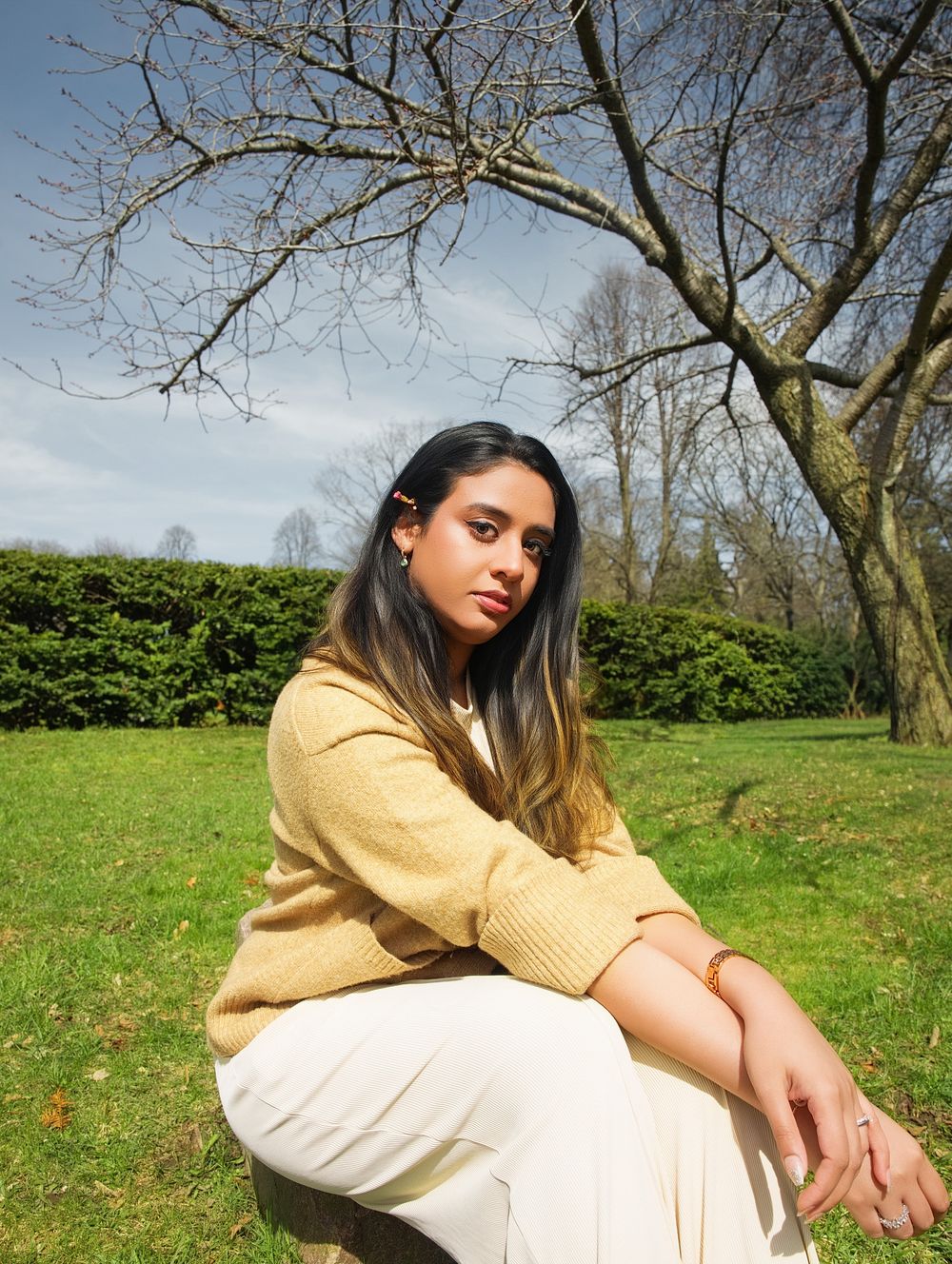 Young woman in yellow sweater and white pants sitting outdoors in a sunny park with green grass and bare trees.