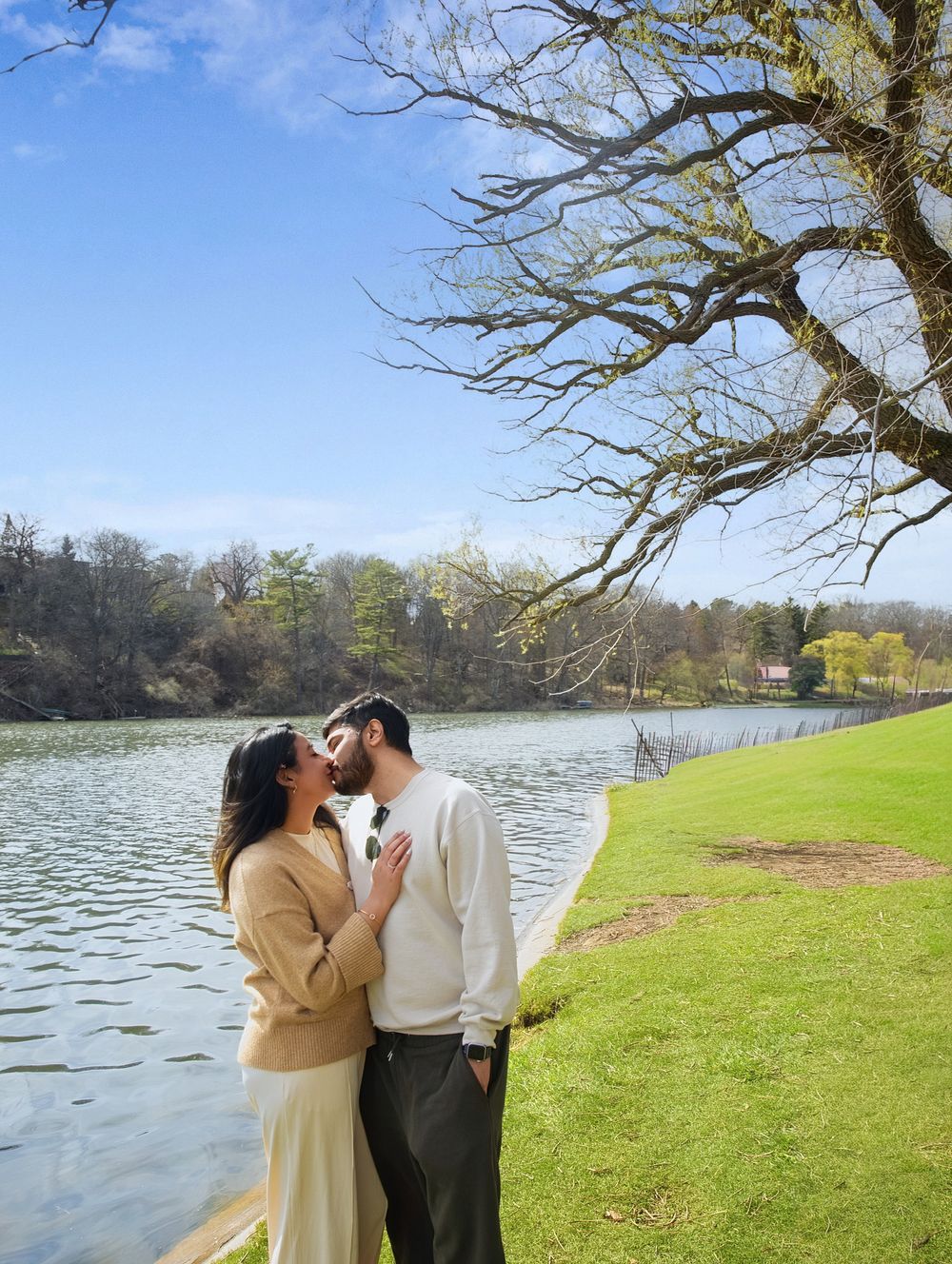 Couple sharing a romantic moment by a serene lake, surrounded by lush greenery and a bare tree under a blue sky.