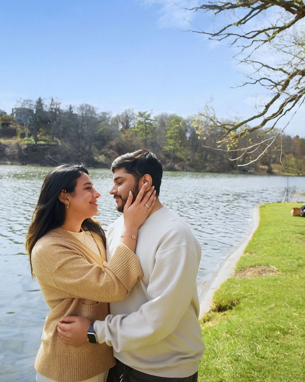 A couple shares a romantic moment by a serene lake on a sunny day, surrounded by lush greenery.