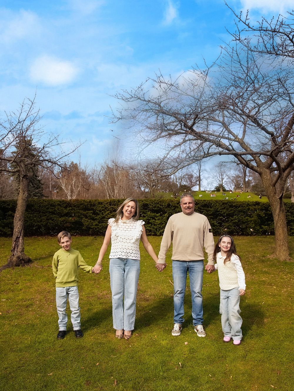 Family of four holding hands and smiling in a green park with bare trees and blue sky.