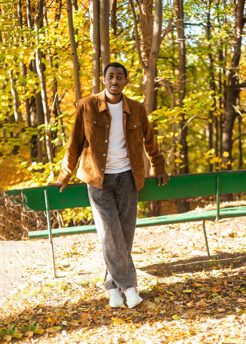 A young Black man walks through an autumn park wearing a brown suede jacket, white tee, and gray jeans.