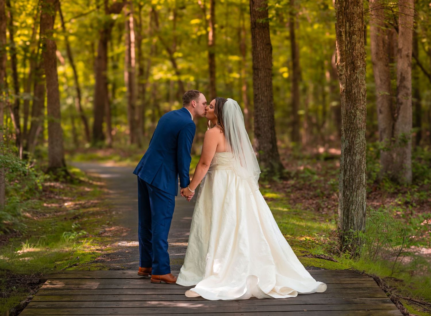 Bride and groom kissing on a wooden path surrounded by lush green forest trees on their wedding day.