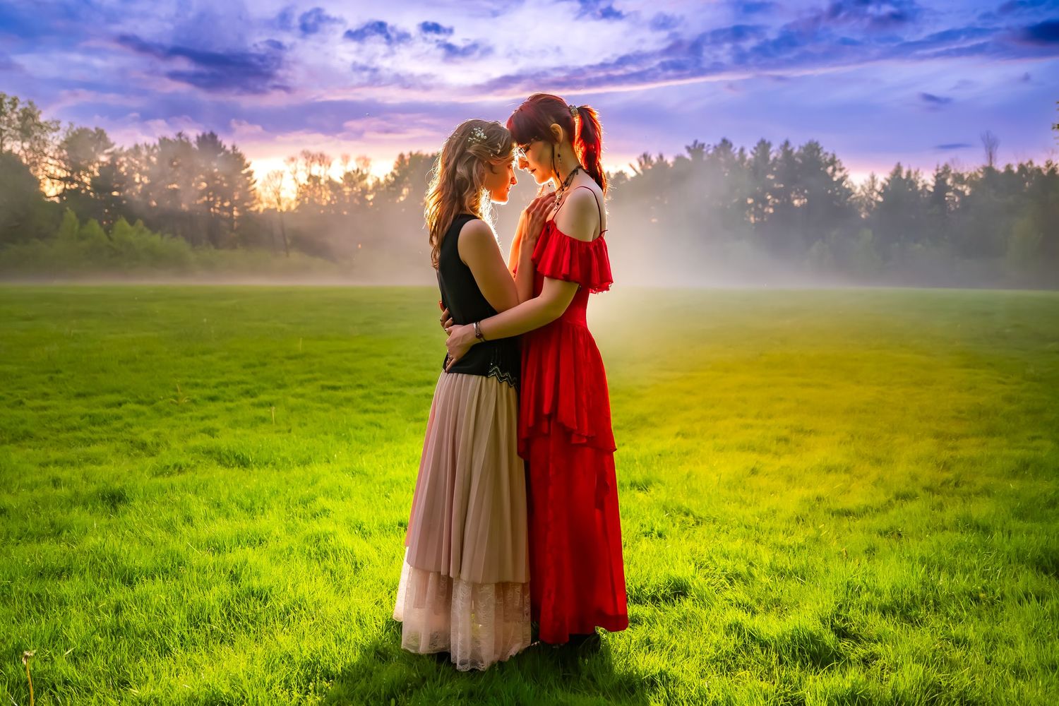 Two women in elegant dresses embrace romantically in a misty green field at sunset with forest backdrop.