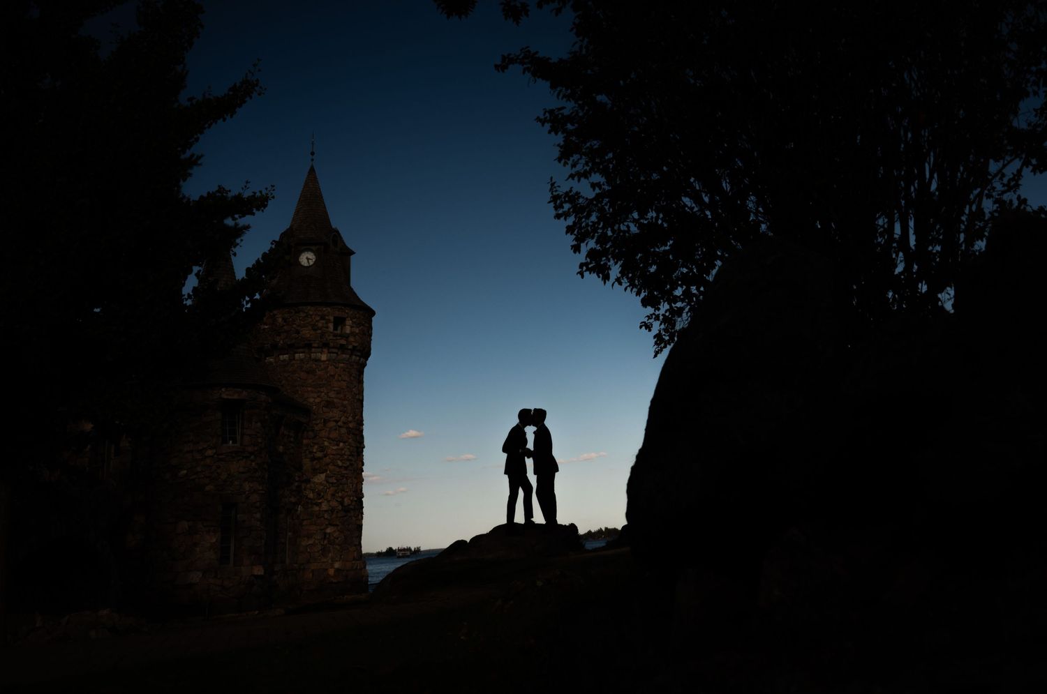 Silhouette of a couple kissing at dusk beside a stone castle tower, framed by trees against a blue twilight sky.