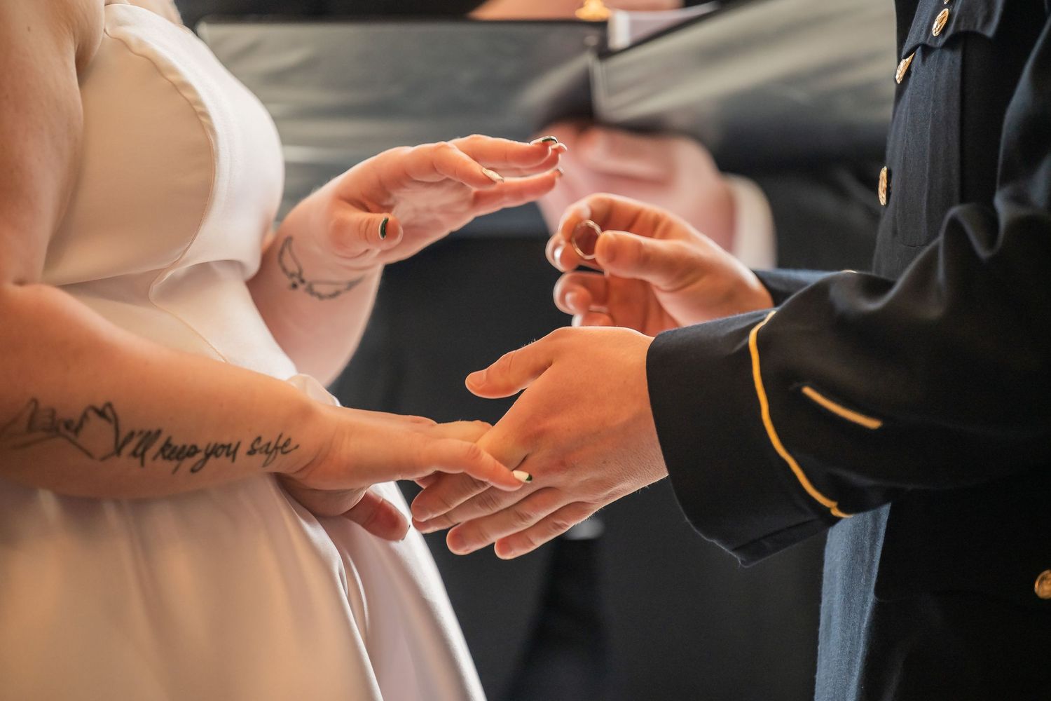 Military groom placing wedding ring on bride's tattooed hand during ceremony.