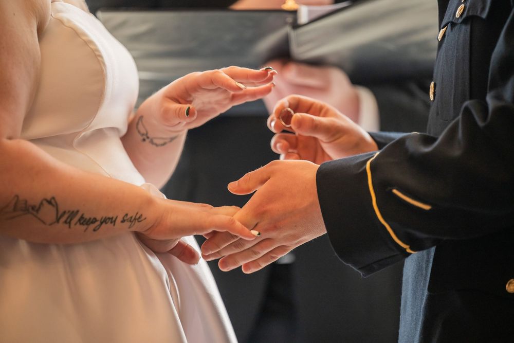Military groom placing wedding ring on bride's tattooed hand during ceremony.