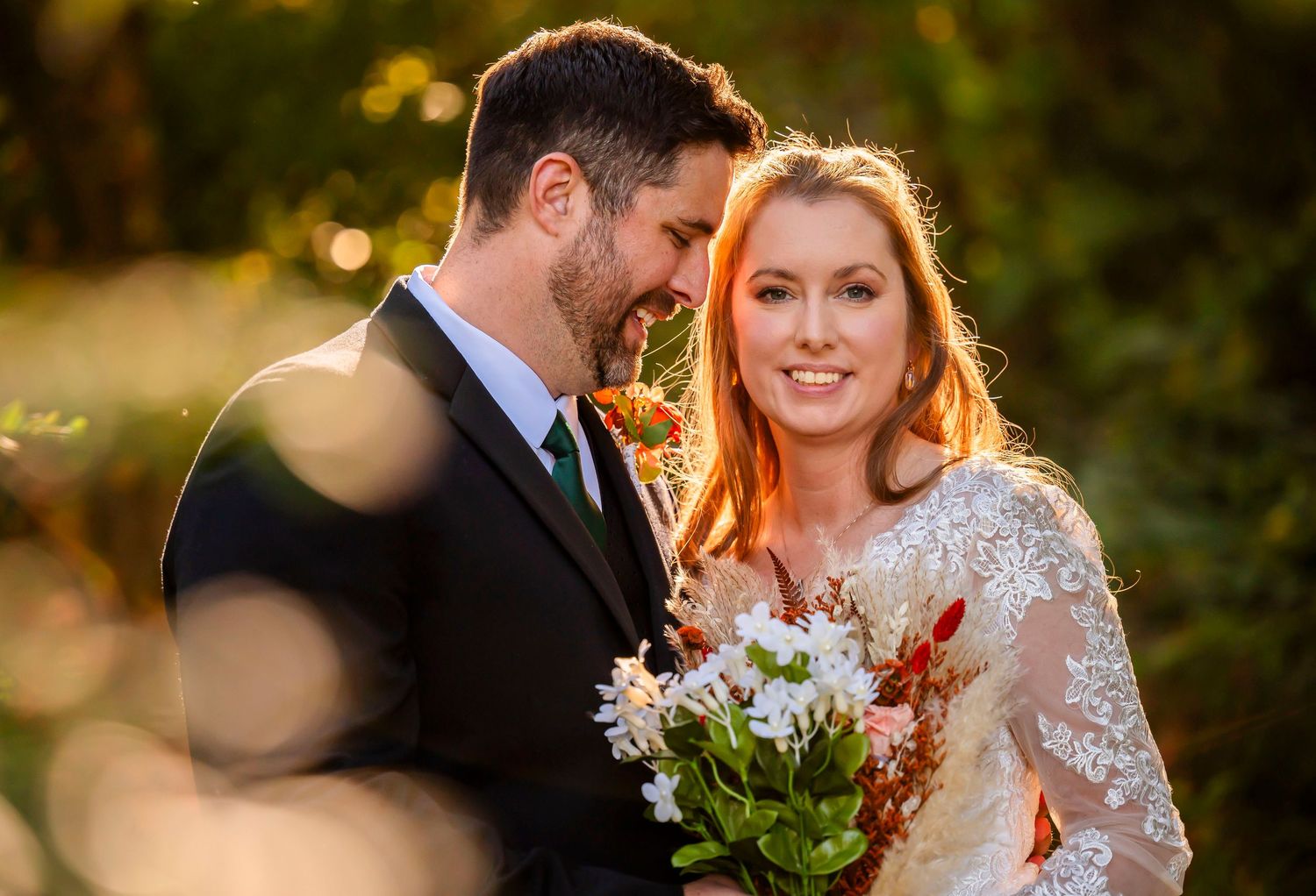 A bride and groom share a romantic moment outdoors, with golden sunlight and lush greenery in the background.