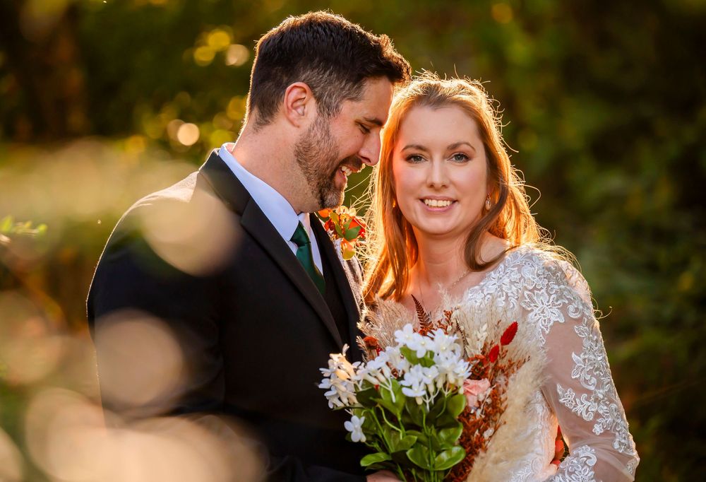 A bride and groom share a romantic moment outdoors, with golden sunlight and lush greenery in the background.