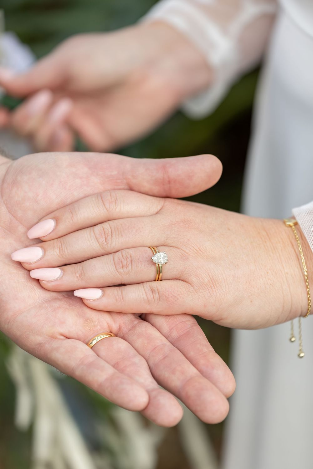 Close-up view of hands wearing wedding rings and delicate jewelry during an outdoor ceremony.