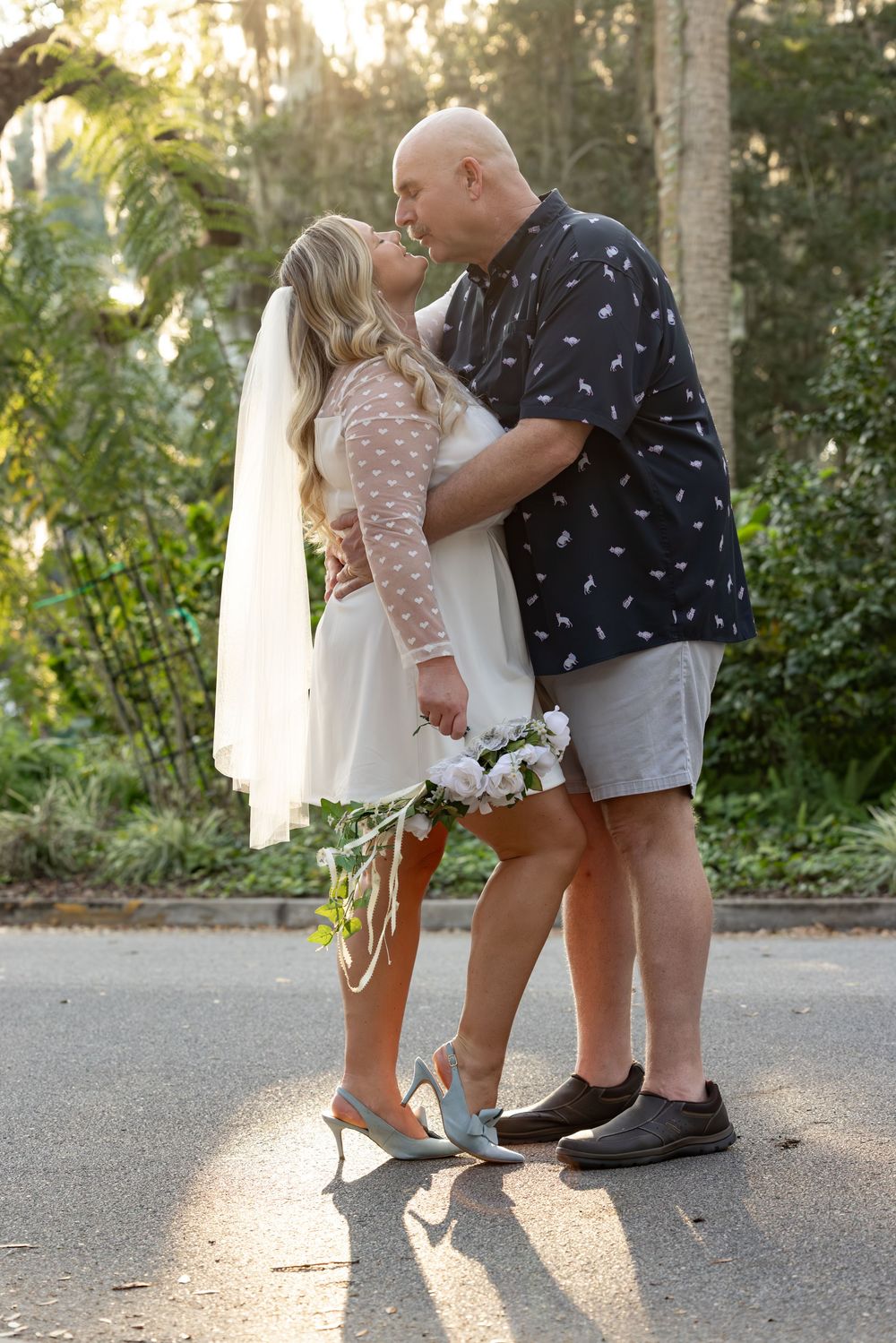 A couple in casual summer attire shares an intimate moment on a sunlit path surrounded by greenery.