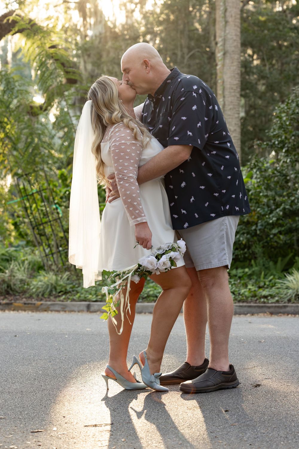 A romantic couple in casual attire sharing tender moments on a sunlit pathway surrounded by greenery.