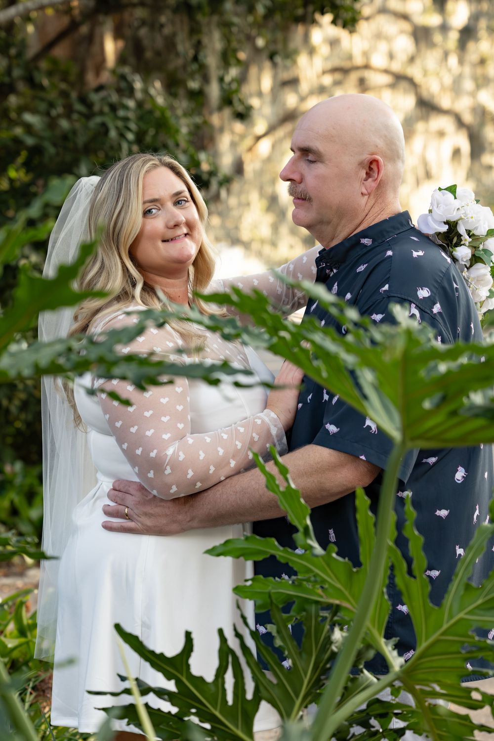 A couple shares a romantic moment in a garden setting with lush tropical plants and dappled sunlight.