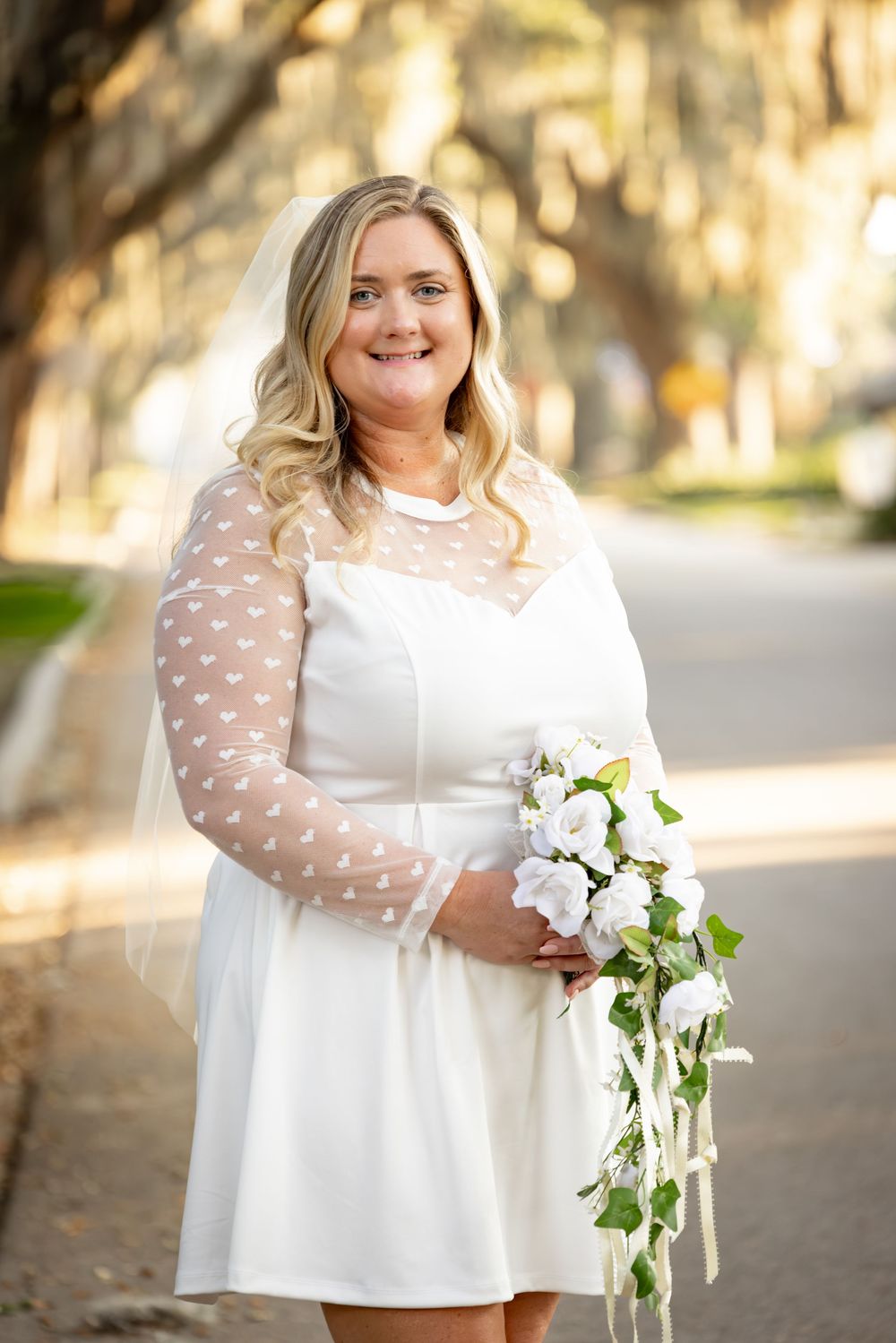 A person in a white dress poses outdoors holding a cascading bouquet of white flowers against a sunlit backdrop.