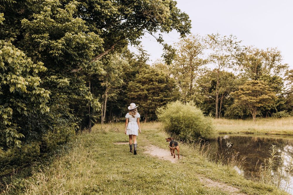 Woman in white dress and cowboy hat walking a dog along a scenic path beside a pond.